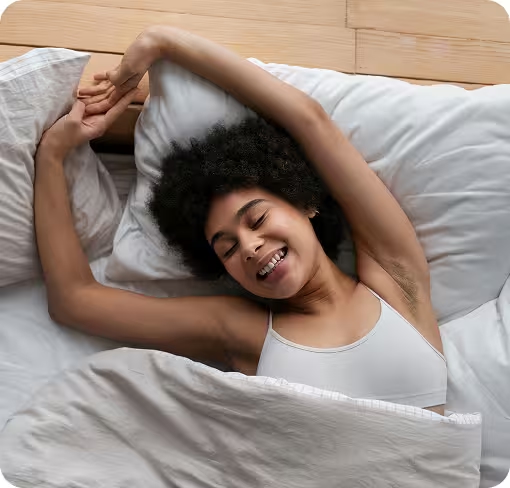 Eine lächelnde junge Frau erwacht ausgeruht im Bett und streckt sich, was guten Schlaf symbolisiert.