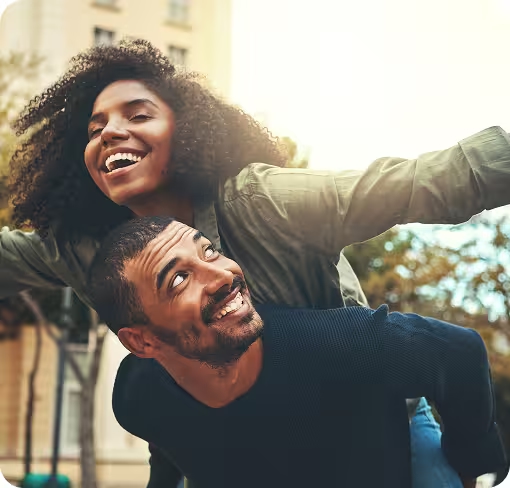 Lachender Mann trägt lachende Frau huckepack, Arme ausgestreckt in der Sonne.