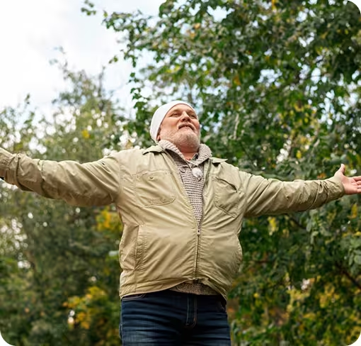 Älterer Mann mit Mütze und Jacke steht lächelnd mit ausgebreiteten Armen im Grünen.
