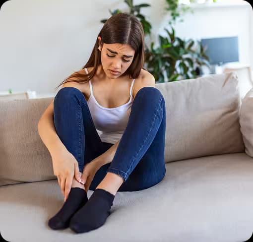 Eine junge Frau sitzt traurig auf dem Sofa und hält ihr Bein, was auf Schmerzen hindeutet.