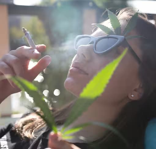 Junge Frau mit Sonnenbrille und Cannabisblättern raucht im Freien einen Joint.