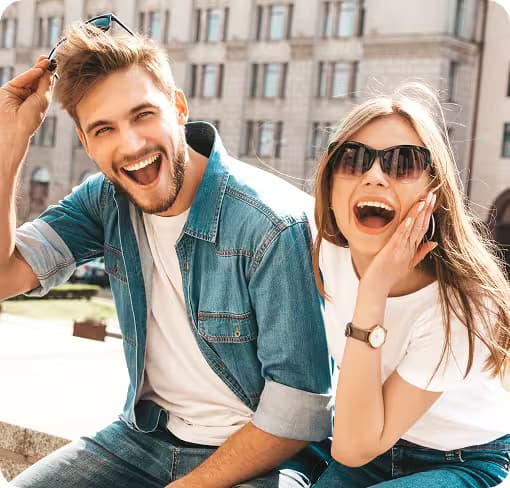 Lachendes, junges Paar in Sonnenbrillen sitzt draußen in der Stadt.